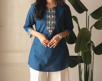 May include: A dark teal tunic featuring three-quarter sleeves and a central vertical panel of floral embroidery in shades of pink, green, and white, paired with wide-leg off-white trousers and pointed-toe neutral-coloured shoes.