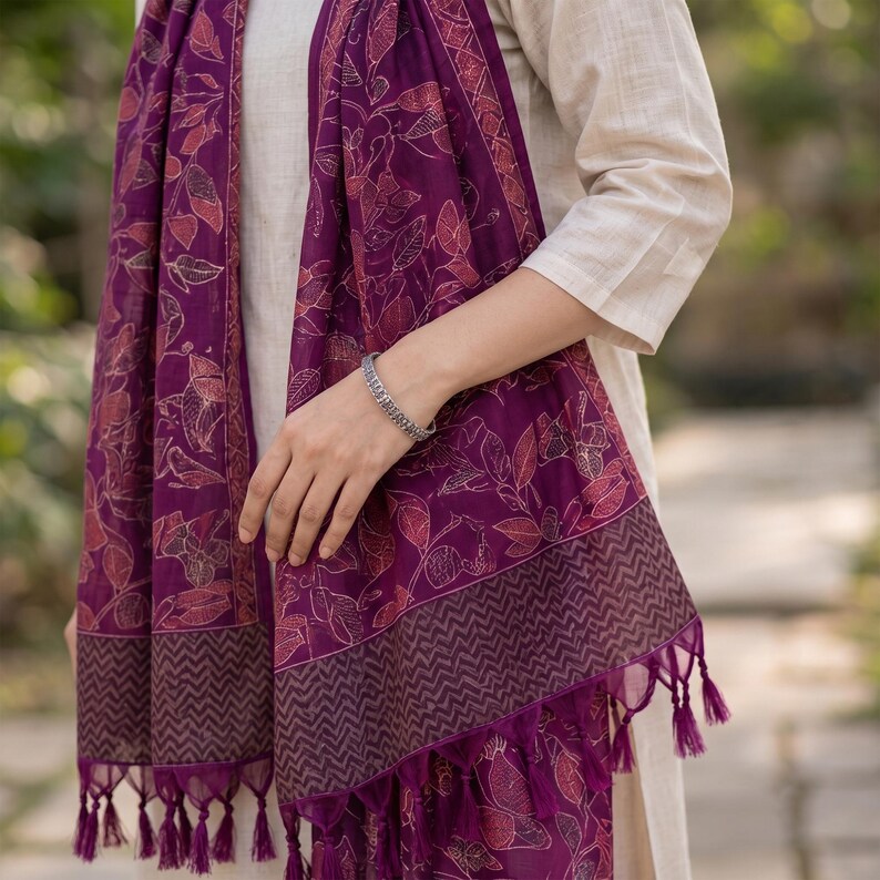 May include: A deep purple scarf featuring a repeating pattern of outlined leaves and vines in a lighter shade. The scarf is finished with a dark chevron-patterned border and decorative tassels along the bottom edge, draped over a light beige garment.