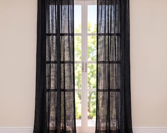 May include: A pair of floor-length, semi-sheer black curtains hanging from a white rod. The fabric has a visible woven texture that allows light to filter through, covering a glass-paned door with a grid frame and silver handles.