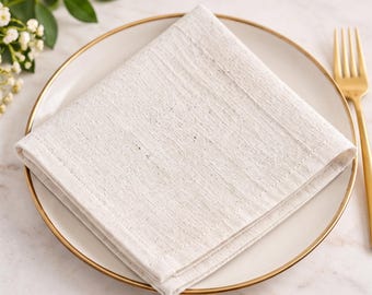 May include: A square, off-white, textured fabric napkin folded into a triangle, placed on a round white plate with a thin gold rim. A gold-toned metal fork rests to the right of the plate, and small white flowers are visible in the corner.