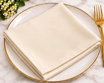 May include: A square, folded cream-coloured cloth napkin centred on a white ceramic plate with a gold-toned rim. The plate features a subtle speckled pattern and is accompanied by a gold-toned metal fork placed to the right side.