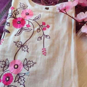 May include: A beige sleeveless tunic featuring embroidered pink flowers with dark purple stems and leaves. The garment has a round neckline and includes the text "Rc kurtis original" printed in the bottom right corner of the fabric.