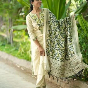May include: A cream-colored three-piece outfit consisting of a long-sleeved tunic with green floral embroidery around the neckline, matching straight-leg trousers with embroidered cuffs, and a large shawl featuring a detailed green leaf print.