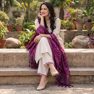 May include: A cream-colored long-sleeved tunic and matching trousers set featuring pink embroidered trim at the hem, paired with a deep purple shawl that has an intricate woven pattern and fringe detailing along the edges, worn with beige heels.