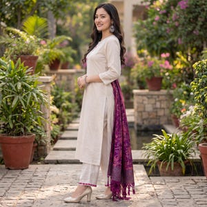 May include: A cream-colored long-sleeved tunic and matching straight-leg trousers featuring a bright pink embroidered hem. A deep purple shawl with a detailed floral pattern and tasseled edges is draped over the side of the outfit.