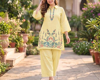 May include: A light yellow two-piece set consisting of a long-sleeved tunic with a collared neckline and wide-leg trousers. The tunic features intricate floral embroidery in shades of pink, green, and blue along the placket and hemline.