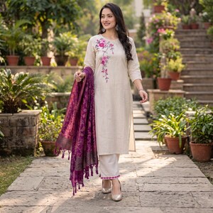 May include: A beige long-sleeved tunic featuring pink and purple floral embroidery on the chest, paired with matching beige trousers that have a pink embroidered hem. A deep purple shawl with a tonal floral pattern and tassels is draped.