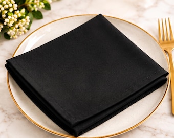 May include: A solid black square cloth napkin folded into a triangle shape, placed centrally on a round white ceramic plate with a thin gold rim, accompanied by a gold-toned metal fork positioned to the right of the plate.