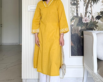 May include: A mustard yellow knee-length tunic with a V-neckline and white trim, featuring three-quarter sleeves with white floral embroidery at the cuffs. The tunic is paired with wide-leg white trousers that have matching white floral embroidery at the hem.