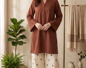 May include: A long-sleeved, solid brown tunic with a V-neckline and side slits, paired with wide-leg, off-white trousers featuring a repeating pattern of small brown hearts. The outfit is completed with simple, light-coloured slide sandals.