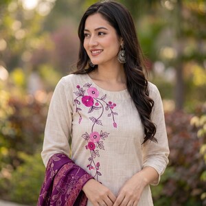 May include: A beige long-sleeved top featuring an embroidered floral vine pattern in shades of pink and magenta. The garment has a round neckline and a textured fabric finish. A deep purple shawl with a gold-toned pattern is draped over one arm.