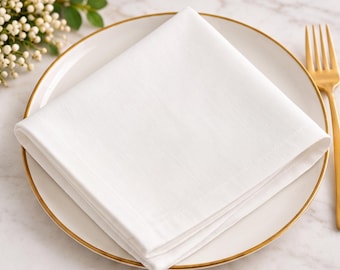 May include: A crisp white square cloth napkin folded neatly and placed on a white ceramic plate with a thin gold rim. A gold-toned metal fork rests to the right of the plate, and a small cluster of white berries is visible in the corner.