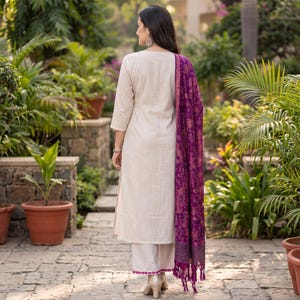 May include: A beige long-sleeved tunic and matching wide-leg trousers featuring a pink embroidered hem, paired with a vibrant purple shawl adorned with an intricate gold floral pattern and finished with long tassels at the bottom edge.