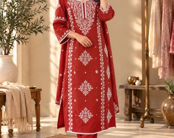 May include: A rust-red long-sleeved tunic and matching trousers set featuring intricate white floral embroidery along the neckline, sleeves, and vertical panels. The outfit includes a coordinating rust-red scarf with white embroidered details.