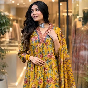 May include: A mustard yellow kurta featuring a floral print in shades of pink, red, and green. The garment has a V-neckline with a decorative border of small mirrors and intricate embroidery, three-quarter sleeves, and a matching yellow dupatta.