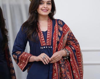 May include: A navy blue kurta featuring a V-neckline with red and white patterned trim, long sleeves with matching cuffs, and a coordinating red, blue, and beige patterned dupatta draped over the shoulder, accented with silver jewellery.