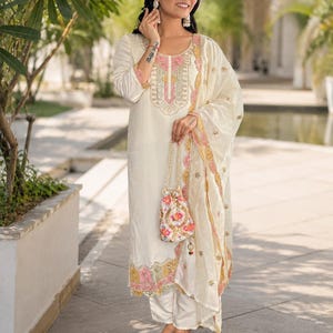 May include: An off-white three-piece outfit consisting of a long-sleeved tunic with pink and gold embroidery, matching straight-leg trousers, and a coordinating dupatta. A small, floral-patterned drawstring pouch with a gold chain strap is included.