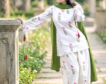 May include: A white long-sleeved tunic and matching wide-leg trousers featuring a repeating pattern of small green stems with pink flower buds. The outfit includes a solid olive green scarf draped over the shoulders and sleeves with tassels.