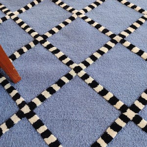 Peut inclure: Un tapis bleu clair orné d'un motif en losanges répétitif. Les lignes de la grille sont composées de segments rectangulaires alternant noir et blanc, créant un design géométrique contrasté sur la surface texturée et unie en bleu.