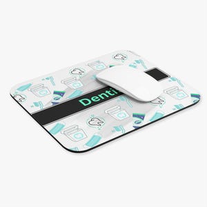 May include: A rectangular mouse pad featuring a white background with a repeating pattern of light blue toothbrushes, dental floss containers, and teeth. A black horizontal band across the center displays the text Denti in light blue.