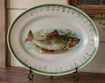 May include: An oval white ceramic platter featuring a central illustration of a brown and green fish swimming among water lilies. The platter has a green rim and a delicate silver filigree pattern along the edge, displayed on a black metal stand.