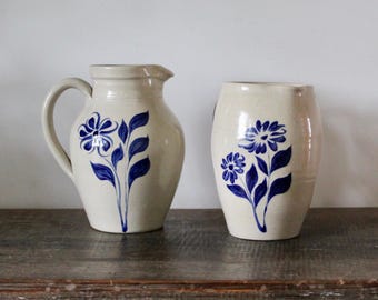 May include: A cream-colored ceramic pitcher with a handle and a matching cream-colored ceramic vase, both featuring hand-painted blue floral designs with petals and leaves on the front surface. The items have a smooth, glossy finish.