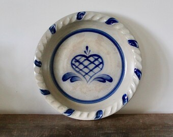May include: A round ceramic pie dish with a light beige base and blue hand-painted details. The center features a blue heart with a cross-hatched pattern, surrounded by a blue ring. The rim has a twisted rope texture with blue accents.