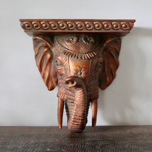 May include: A wall-mounted shelf featuring a sculpted elephant head in a metallic bronze finish. The shelf top has a decorative rope-like border, while the elephant head displays intricate floral carvings on the forehead and textured skin details.