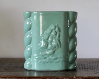 May include: A light green ceramic vase featuring a raised relief of a sailing ship on the front panel. The sides of the vase are decorated with a vertical rope-like texture, and the entire piece has a smooth, glossy finish.