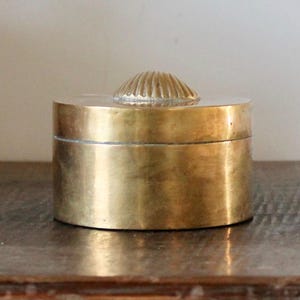 May include: A cylindrical brass box with a polished metallic finish. The lid features a centered, raised, dome-shaped handle with a textured, radiating ribbed pattern resembling a seashell. The container has a smooth, uniform side profile.