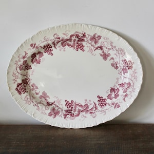May include: An oval-shaped ceramic serving platter with a cream-colored base and a decorative border featuring deep red illustrations of grape clusters and vine leaves. The platter has a scalloped, fluted edge design around the entire perimeter.