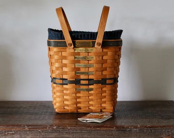 May include: A rectangular woven wood basket with two light brown vertical handles. The basket features horizontal dark green woven bands and several small brass-colored rectangular plaques attached to the front. A dark blue fabric liner is visible.