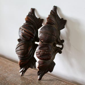 May include: A pair of dark brown carved wooden wall ornaments featuring detailed depictions of rounded fruits or nuts nestled among textured leaves. The wood displays a rich, deep grain with intricate cross-hatched patterns on the surfaces.