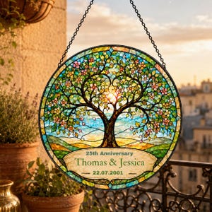 Op de afbeelding: Een ronde glas-in-lood zonnevanger met een boom met groene bladeren en roze bloesems. Een centraal lint toont de tekst 25th Anniversary, Thomas & Jessica en 22.07.2001. Het object hangt aan een zwarte metalen ketting.