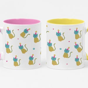 May include: Two white ceramic mugs featuring a repeating pattern of teal cats with yellow striped bodies wearing pink party hats. One mug has a pink handle and interior, while the other mug has a yellow handle and interior with small hearts and flowers.