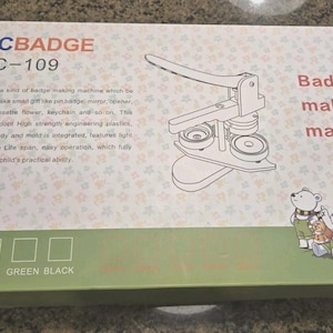 May include: A rectangular product box for a DCBADGE DC-109 badge making machine. The white box features a line drawing of the machine, the text Badge making machine, and a group of cartoon bears. A green bottom panel lists color options.