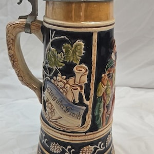 May include: A ceramic beer stein with a dark blue body, featuring a metal lid and a handle shaped like a tree branch. The stein displays embossed relief designs of grapevines, a scroll with the text "Ein guter Trunk", and a figure in traditional attire.