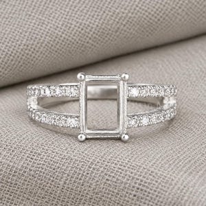 May include: A silver-toned ring featuring a rectangular center setting designed for a gemstone, flanked by a split shank band. The band is adorned with small, clear pavé-set stones along both sides, creating a bright, metallic appearance.