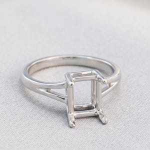 May include: A silver-toned metal ring setting featuring a rectangular prong head designed to hold a gemstone. The band has a split shank design that tapers toward the center setting, creating a polished and clean look for a custom jewelry piece.