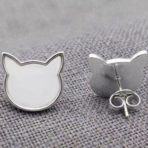 May include: A pair of silver-toned stud earrings shaped like cat heads. Each earring features a smooth, white enamel inlay within a polished silver-colored metal border. One earring is shown from the front, while the other shows the back post.