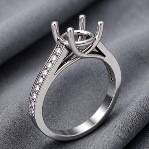 Könnte beinhalten: Eine silberfarbene Ringfassung aus Metall mit einem vierzinkigen Kopf zur Aufnahme eines Mittelsteins. Der Ring ist mit einer Reihe kleiner, runder, klarer Steine besetzt, die entlang der Ringschiene eingelassen sind und für ein glänzendes Aussehen sorgen.