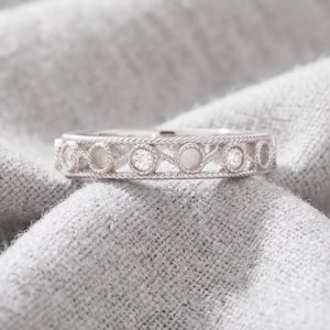 Puede incluir: Un anillo de tono plateado con un patrón repetitivo de engastes circulares conectados por bandas metálicas trenzadas. El diseño alterna entre pequeñas piedras redondas transparentes y marcos circulares vacíos a lo largo del anillo.