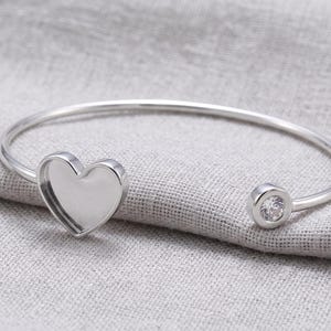 May include: A silver-toned open cuff bracelet featuring a heart-shaped bezel setting on one end and a single round clear stone set in a circular bezel on the opposite end. The metal has a polished finish and a smooth, curved band design.