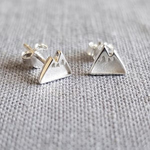 Könnte beinhalten: Ein Paar silberne Ohrstecker in Form von Berggipfeln. Jeder Ohrstecker hat eine dreieckige Silhouette mit einer texturierten, matten Oberfläche im unteren Bereich und einer glatten, polierten Silberkontur für die Schneekappen.