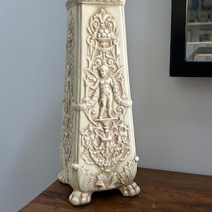 May include: A tall, off-white ceramic obelisk vase featuring intricate relief carvings of winged cherubs, floral garlands, and ornate scrollwork. The piece has a crackled glaze finish and stands on four small, decorative claw-style feet.