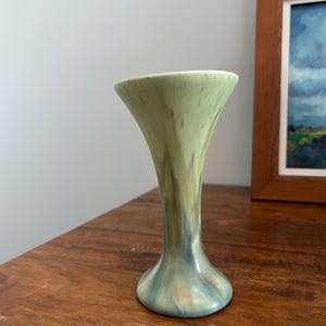 May include: A ceramic vase with a flared rim and a narrow neck that widens at the base. The surface features a mottled glaze in shades of sage green, soft blue, and warm brown, creating a blended, flowing appearance across the entire body.