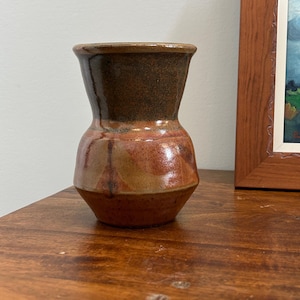 Peut inclure: Un petit vase en céramique avec un bord évasé et un col effilé. La partie supérieure présente une glaçure tachetée brun foncé, tandis que le corps inférieur, plus large, affiche un mélange de tons terre cuite, beige et brun rougeâtre.