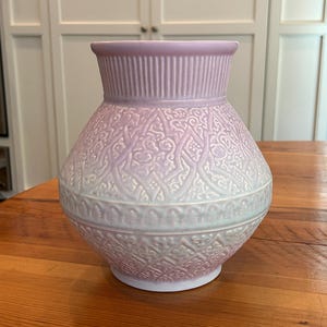 May include: A ceramic vase featuring a soft lavender and white ombre color gradient. The neck has vertical ribbing, while the rounded body displays an intricate, embossed geometric pattern with swirling motifs throughout the surface.