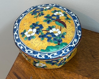 May include: A round ceramic lidded box featuring a vibrant yellow background with a repeating pattern of blue and white flowers, green foliage, and a small bird. The lid and base are bordered by a dark blue rim with a white scrolling vine design.