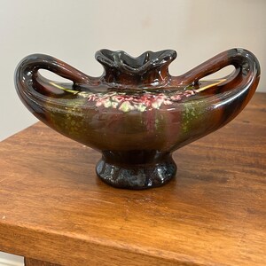 May include: A ceramic vase with two handles and a ruffled rim, featuring a glossy glaze in shades of dark brown, olive green, and deep red. The body of the vase is decorated with a raised, textured floral relief pattern in pink and green tones.